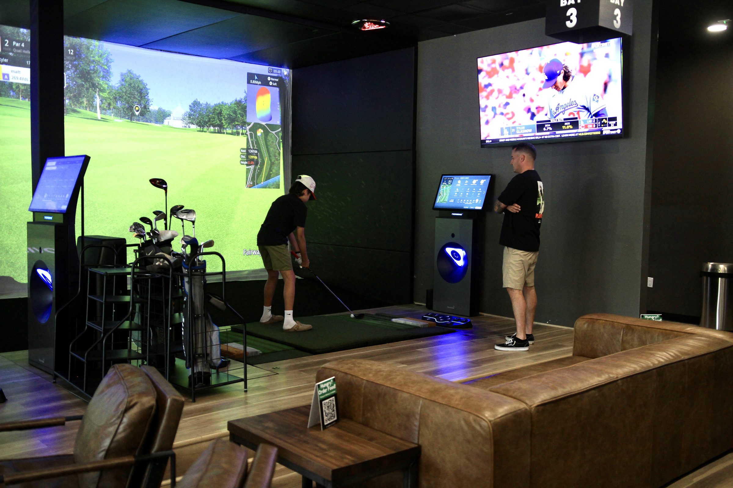 A player hitting in a simulator bay while a friend watches, a ball game on the TV above and leather lounge seating in front.